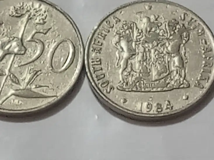 Two 50 cent coins, one showing a number '50' and the other with coat of arms  and  mintage '1984' comprising dual language text, on a reflective surface.