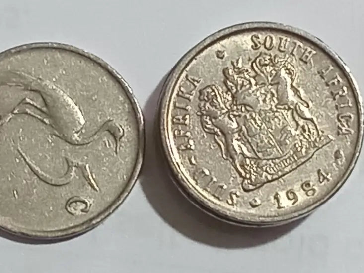 Two 1984 5 cent coins from South Africa displaying a coat of arms and  the other with the denomination on a light gray background.