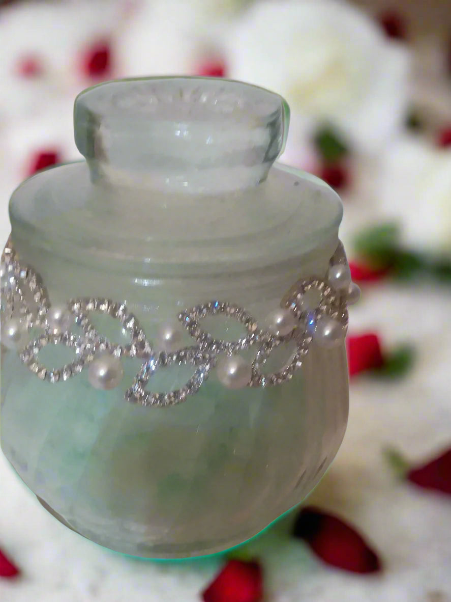 Clear glass jar filled with teal soy granules with decorative leaf chain and pearls on a white band. 